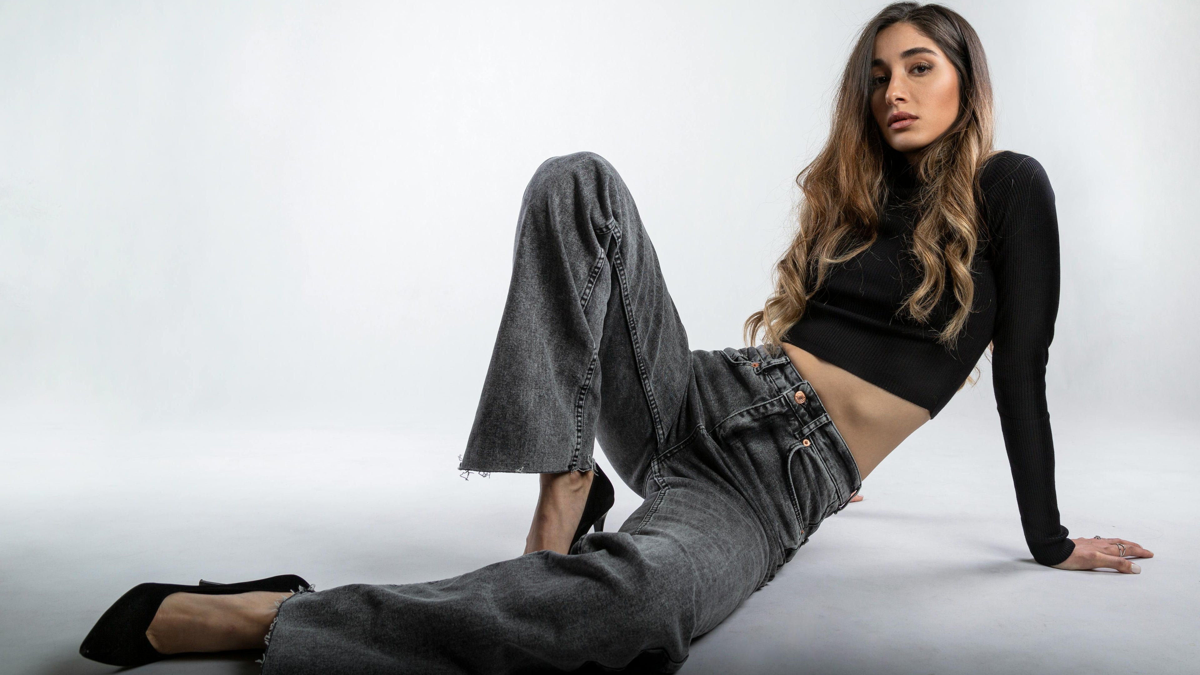 Woman sitting on the floor wearing a black top and gray jeans against a white background